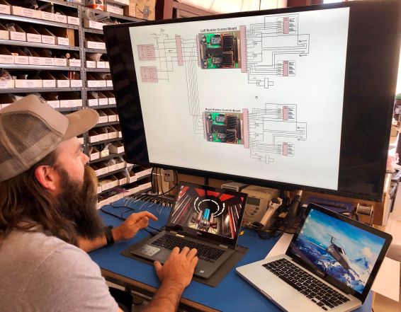 a man with a big beard and baseball cap is working on two small laptops hooked up to a big monitor with a wiring diagram