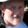 Close-up portrait of an older Caucasian man with light-colored skin, graying hair, and a short white beard, wearing a hat and a light-colored shirt.