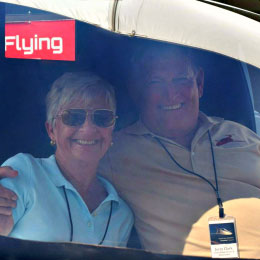 An older couple smiles for a photo while seated together in an airplane cockpit; the woman wears sunglasses and a light blue polo shirt, and the man wears a tan polo shirt with a lanyard and ID badge hanging around his neck.