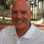 A middle-aged man with a shaved head and a light complexion, wearing a white collared shirt, smiles while looking directly at the camera.