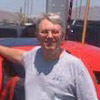 A middle-aged man with short, light hair and glasses wears a light-colored sleeveless shirt, standing outdoors with a bright red vehicle partially visible in the foreground.