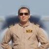 A man with short brown hair wearing sunglasses and a tan military flight suit, standing in front of a blurred aircraft.