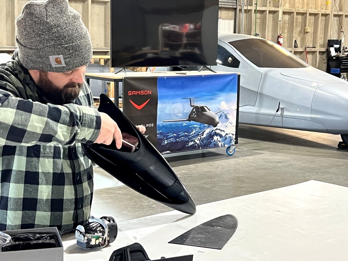 Tanner Making Headlight A bearded man wearing a grey beanie and plaid shirt works on assembling a black, aerodynamic automotive part, likely a headlight housing, at a table in a workshop, with a partial flying car prototype and a promotional banner for a "Samson Switchblade" visible in the background.