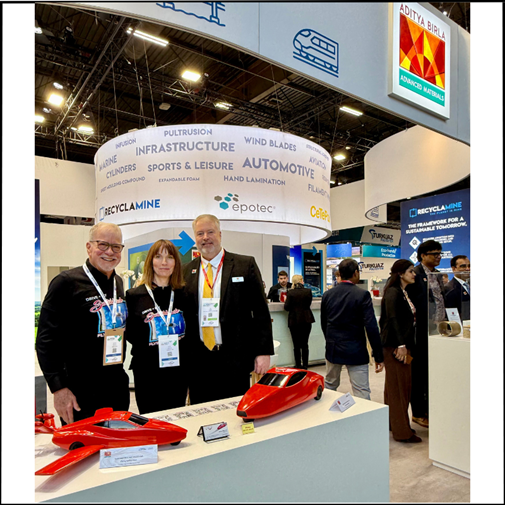 Exhibits_1 Three people standing at an Aditya Birla Advanced Materials trade show booth, which features hanging circular signage with labels like "Infrastructure," "Automotive," and "Wind Blades." Two small, red car-shaped prototypes are displayed on a white counter in the foreground, while other attendees are visible in the background.